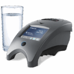 A sleek, modern WaterLink Spin Touch DW - 3585 water testing device with a touchscreen display sits next to a clear glass of water on a white surface. The device shows testing options on its screen.