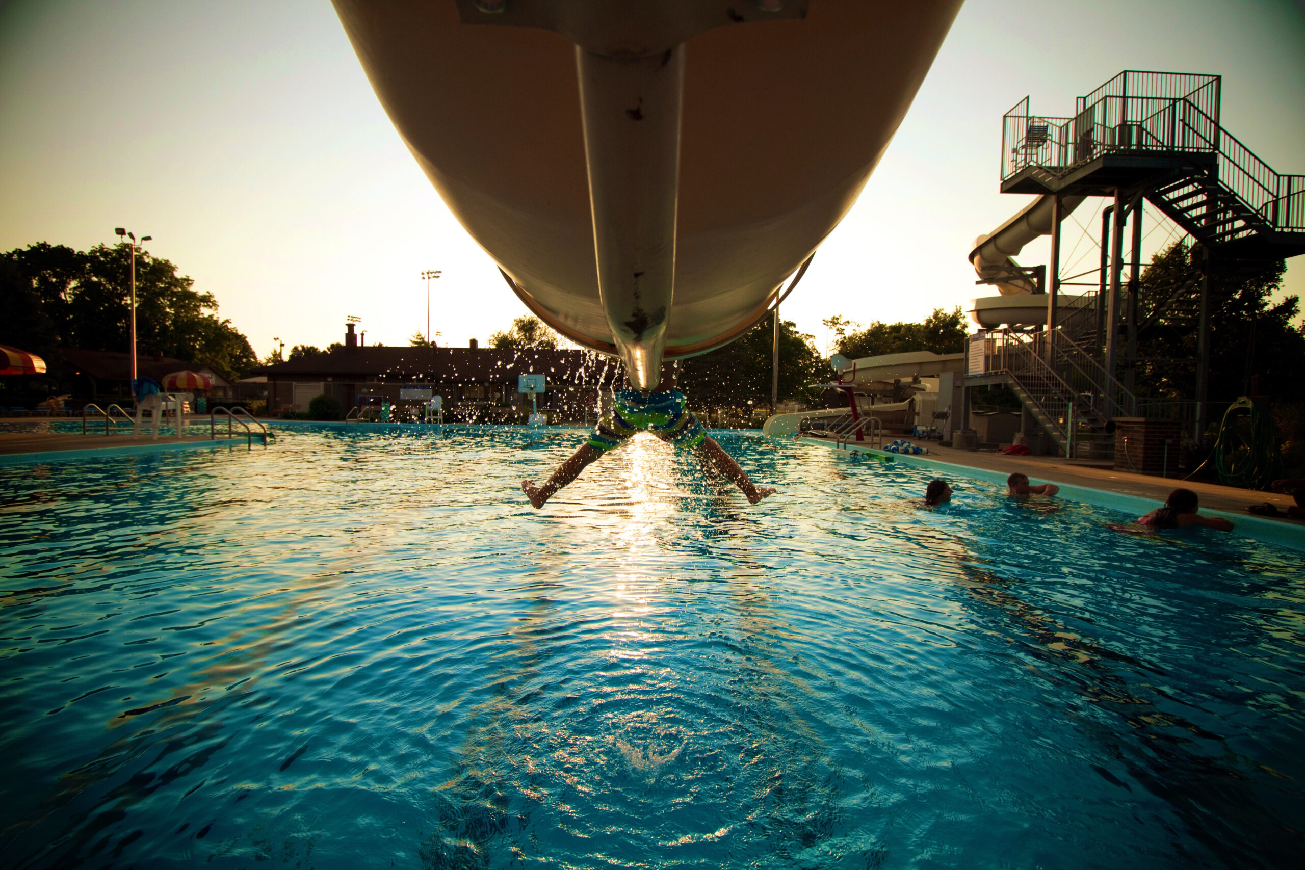 A person comes down a waterslide and splashes into a swimming pool, with water droplets in midair and other swimmers nearby. The sun is setting, casting a warm glow over the scene.
