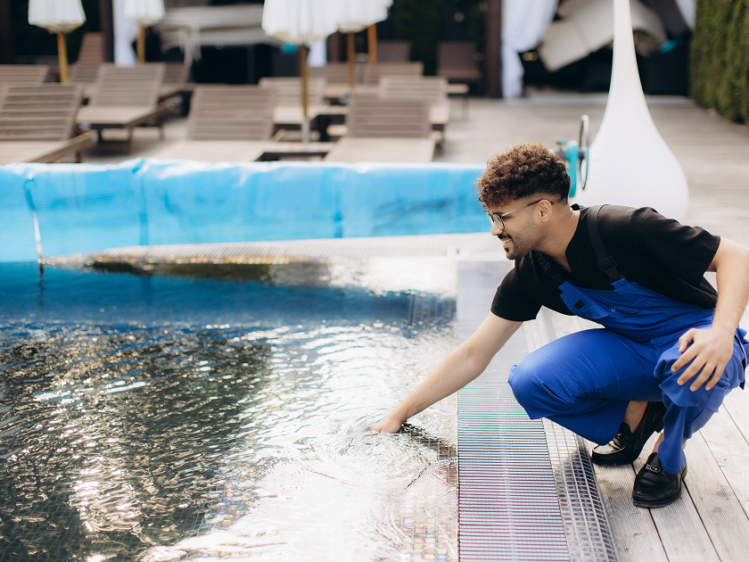 Un hombre con mono azul y gafas se agacha junto a una piscina, metiendo la mano en el agua. En el fondo se pueden ver tumbonas y sombrillas en una terraza de madera. 
